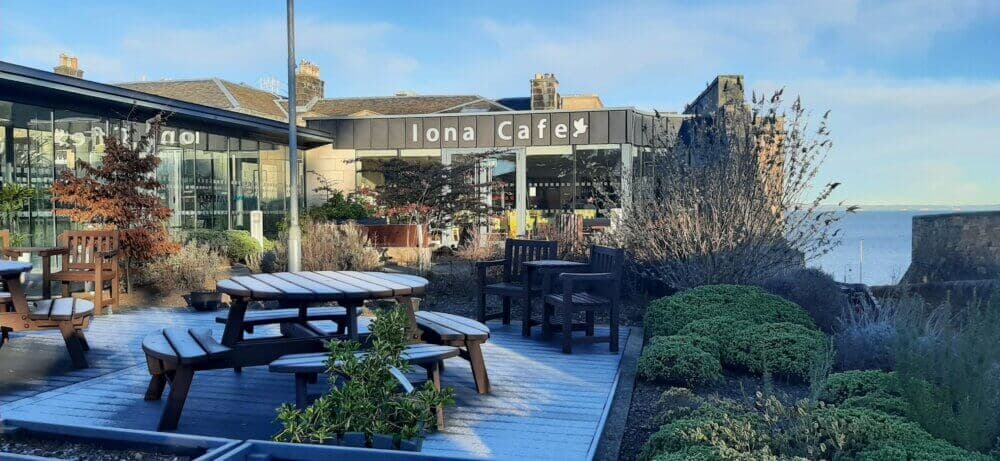Outdoor seating area of Ilona Cafe with wooden benches and tables, situated near the sea. - Home Instead