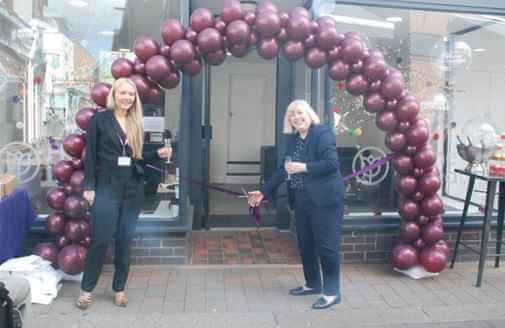 Two people stand in front of a balloon archway, holding a ribbon-cutting ceremony outside a storefront. - Home Instead