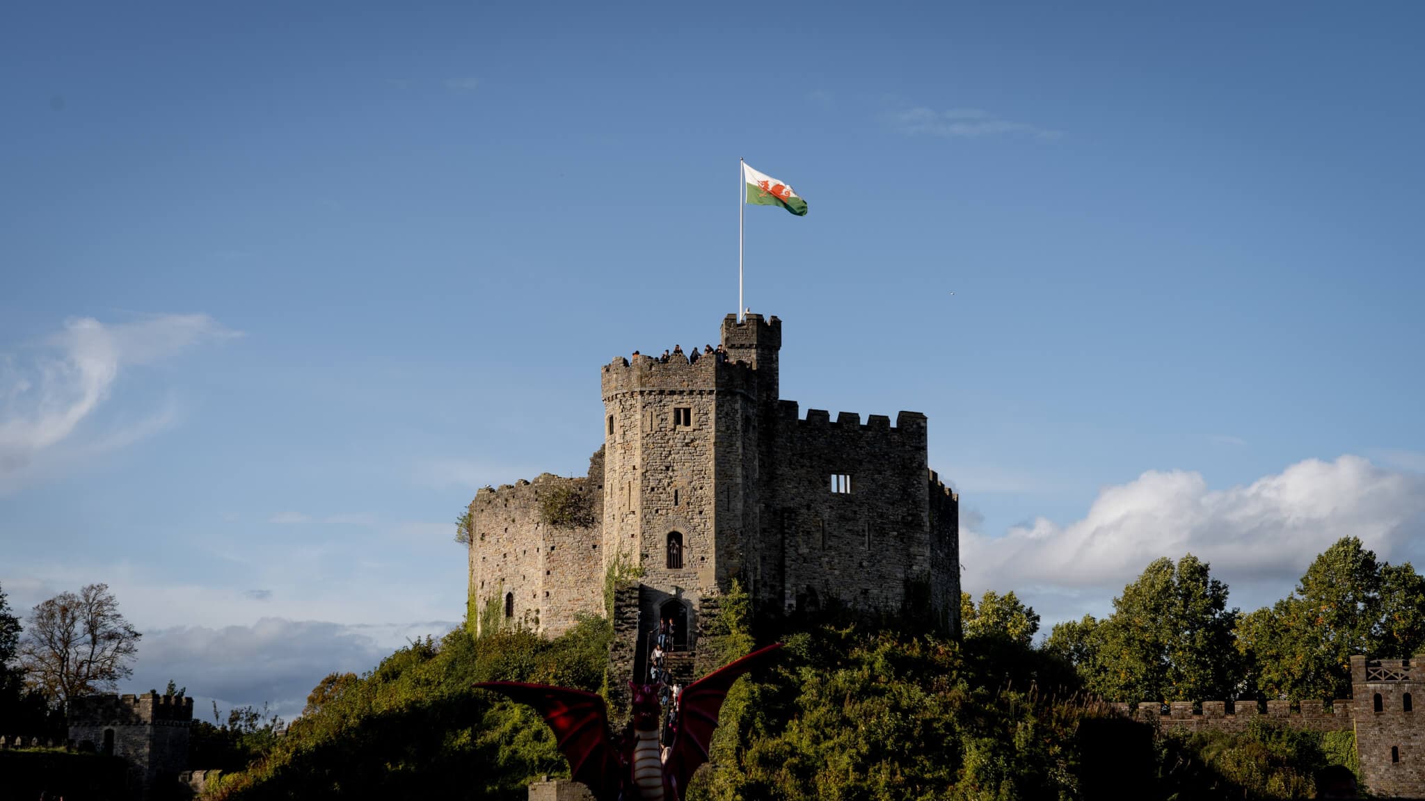 Stone castle with a Welsh flag flying on top, surrounded by greenery under a blue sky with a dragon sculpture in front. - Home Instead