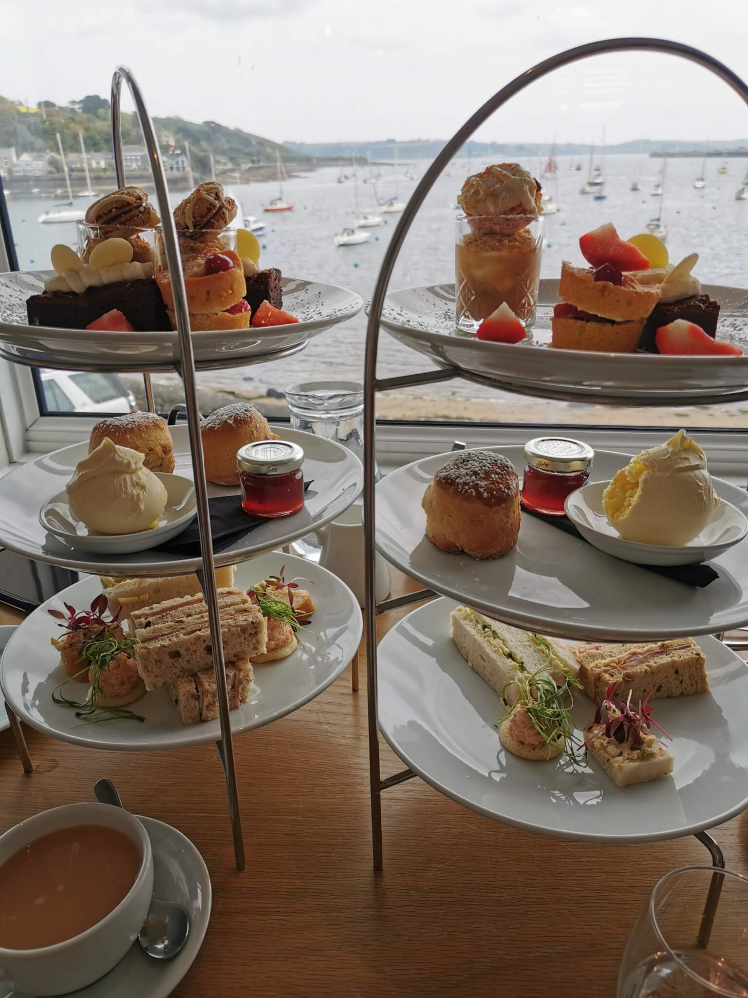 Afternoon tea setup with cakes, scones, and sandwiches on a three-tier stand by a window overlooking a marina. - Home Instead