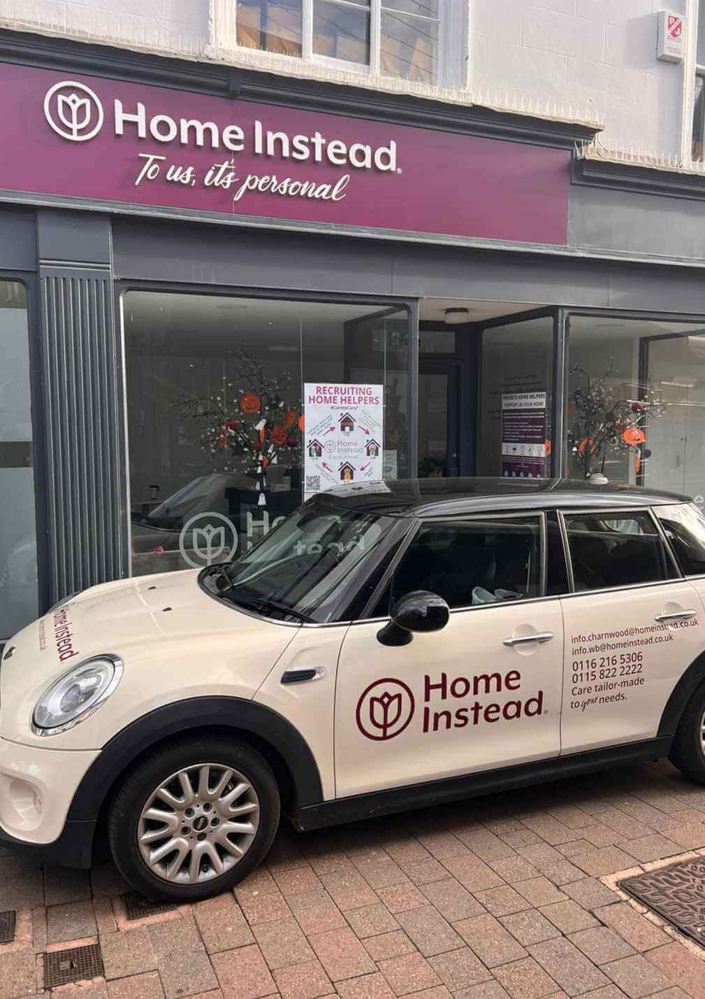 Home Instead office with branded car parked outside, displaying recruitment poster and festive decorations in the window. - Home Instead