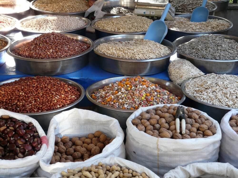Large metal bowls and sacks filled with various nuts, seeds, and grains displayed at a market stall. - Home Instead