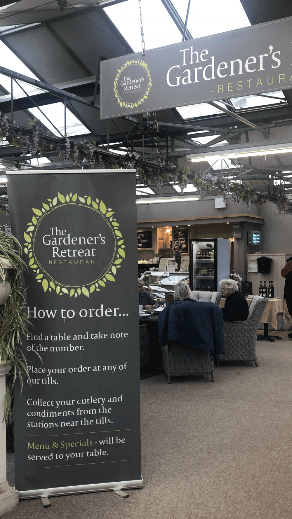 Banner and seating area inside The Gardener's Retreat restaurant, with instructions on how to place and collect orders. - Home Instead