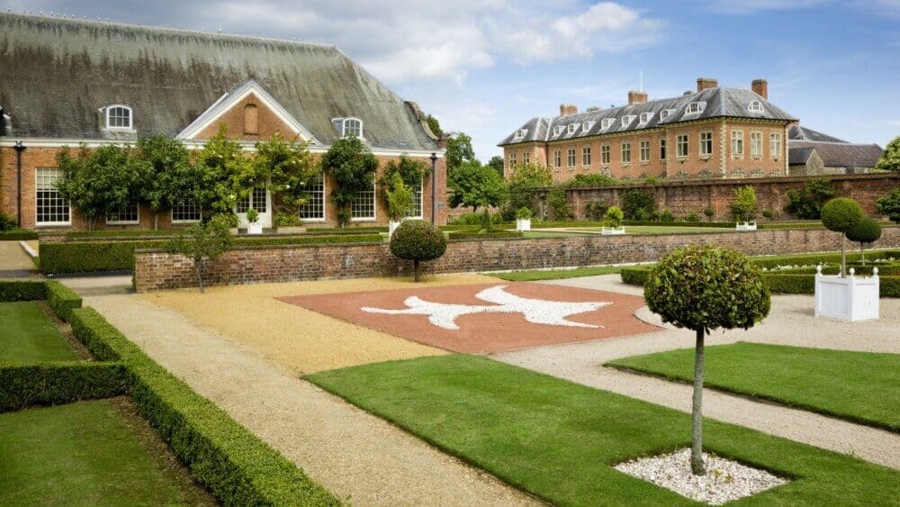 Manicured gardens with hedges, paths, and topiary trees in front of classic brick buildings under a blue sky. - Home Instead