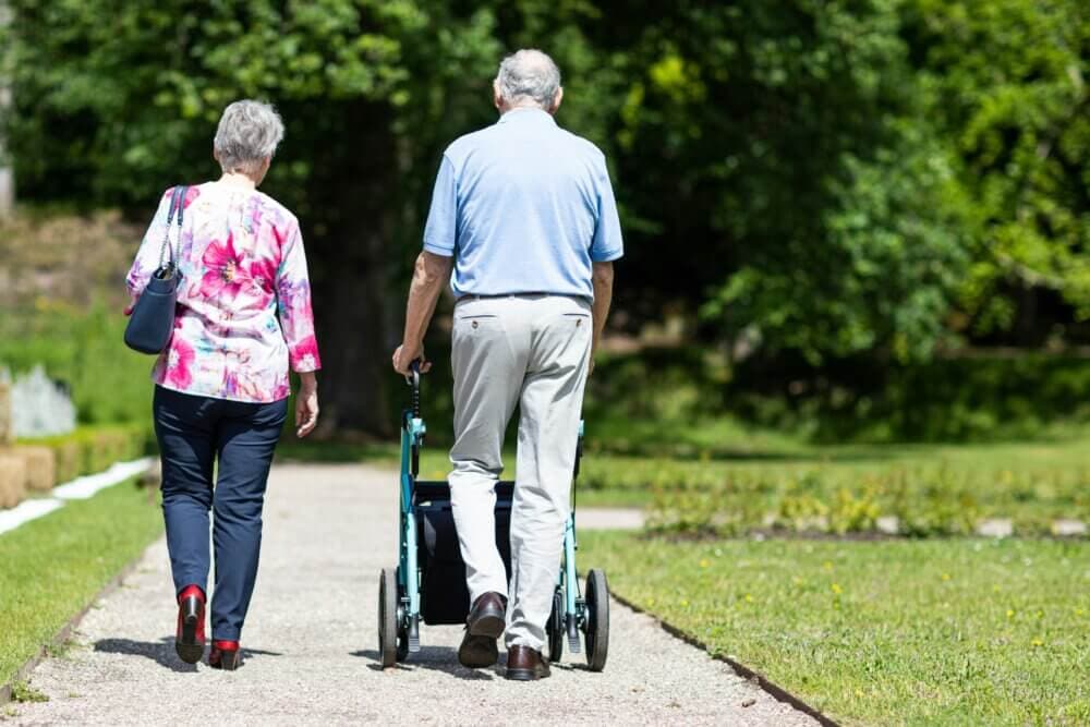 An elderly couple walking in a park, the man using a walker and the woman carrying a handbag. - Home Instead