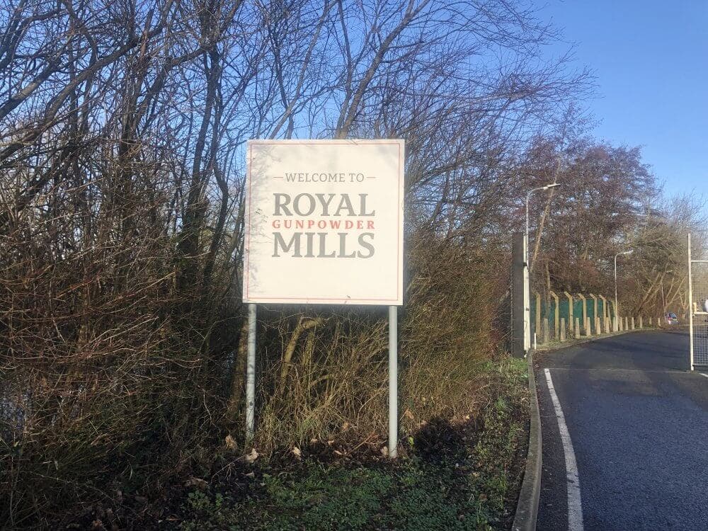 Sign reading "Welcome to Royal Gunpowder Mills" on a roadside with trees and fence in the background on a sunny day. - Home Instead