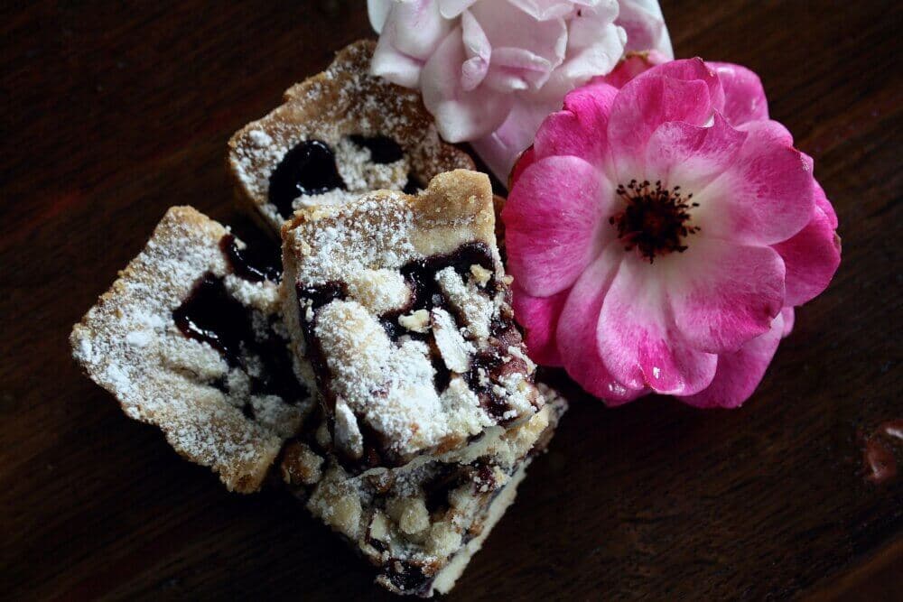Four crumble-topped berry bars dusted with powdered sugar, placed next to a pink and white flower on a dark wooden surface. - Home Instead