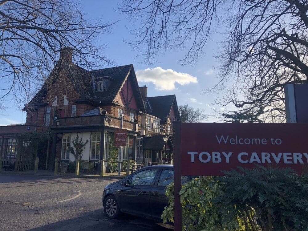 A Toby Carvery restaurant on a sunny day with a black car parked outside and trees in the background. - Home Instead