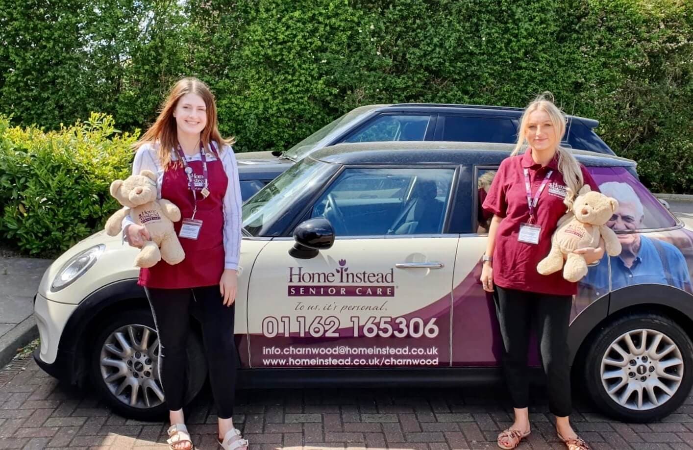 Two women holding teddy bears stand in front of a car with "Home Instead Senior Care" branding. - Home Instead
