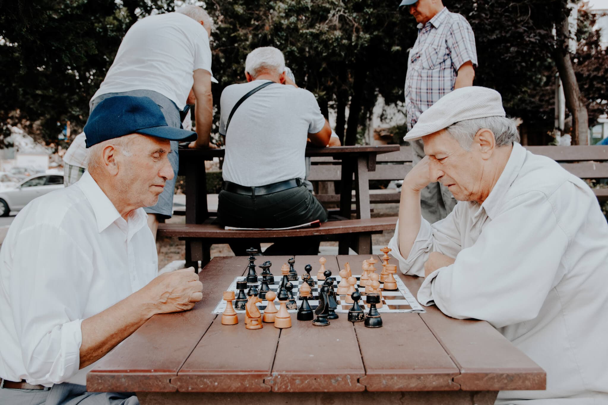 Two elderly men playing chess outdoors on a wooden table, with other people in the background. - Home Instead