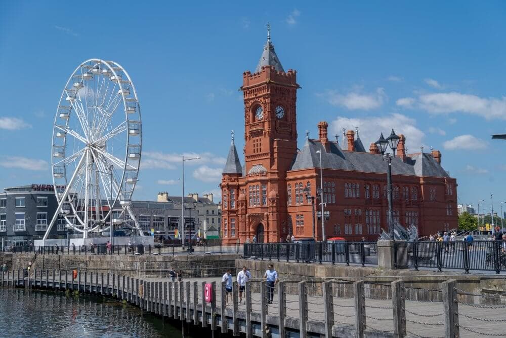 A large Ferris wheel and a historic red-brick building by a waterfront under a blue sky with scattered clouds. - Home Instead