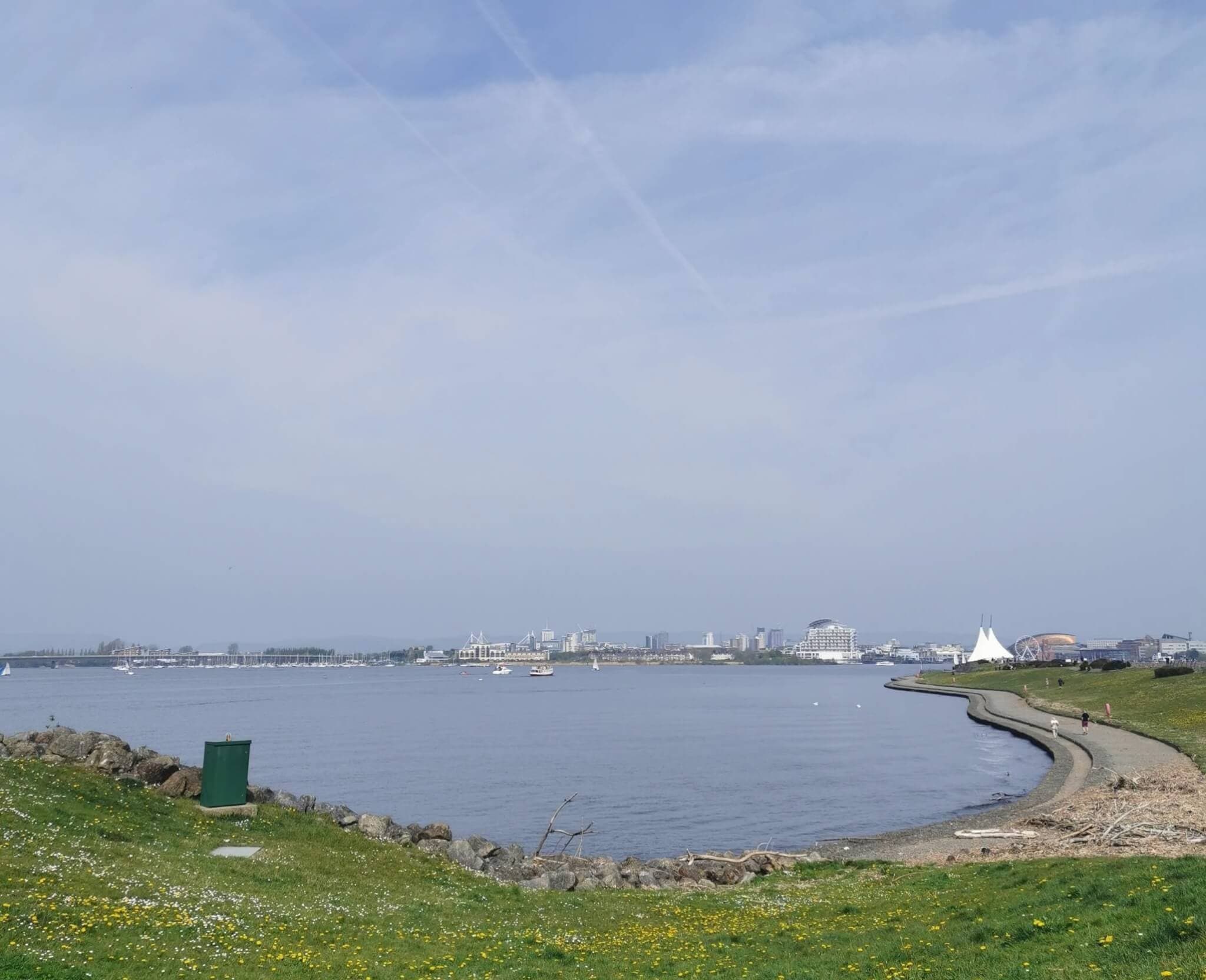 A paved path curves along a calm waterfront with distant city buildings and clear skies above. - Home Instead