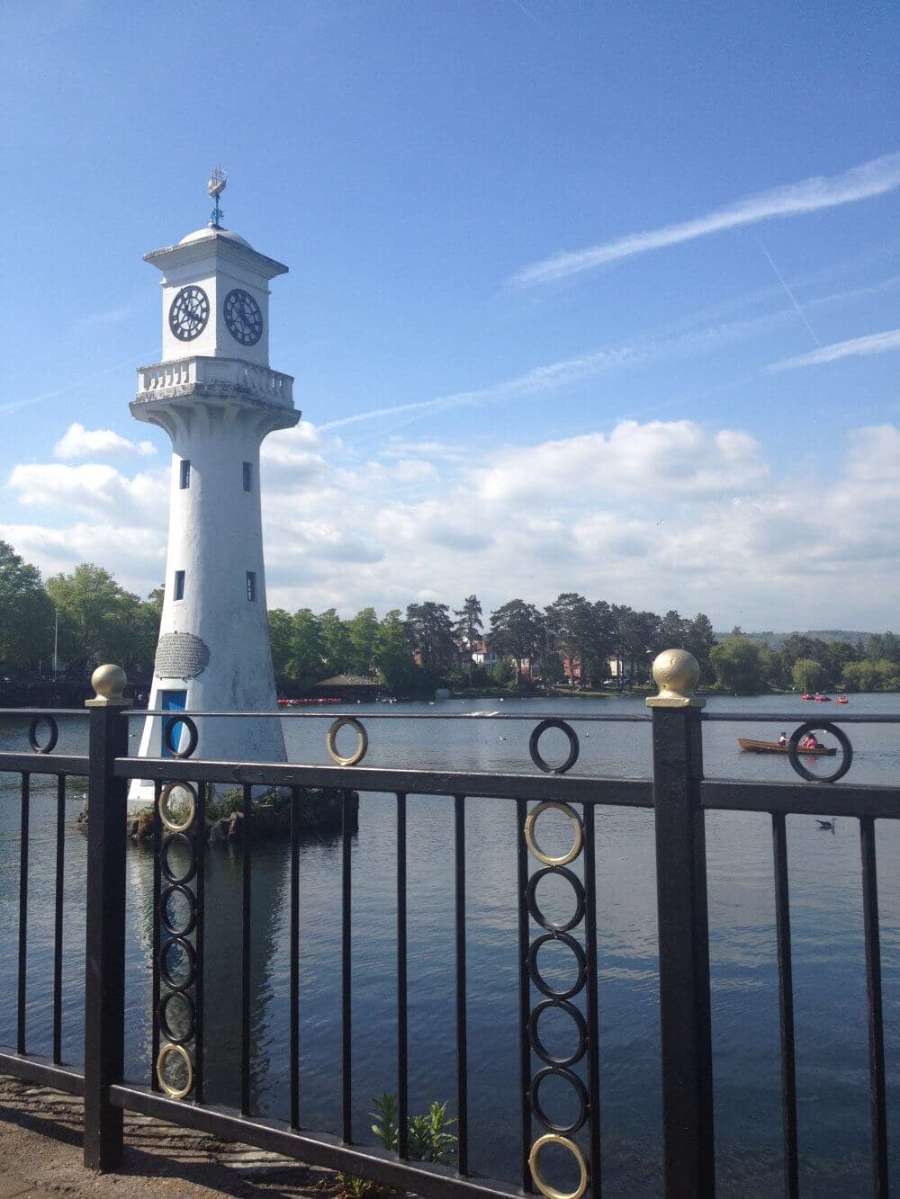 A white clock tower stands by a calm lake, fenced in by a railing, under a partly cloudy blue sky. - Home Instead