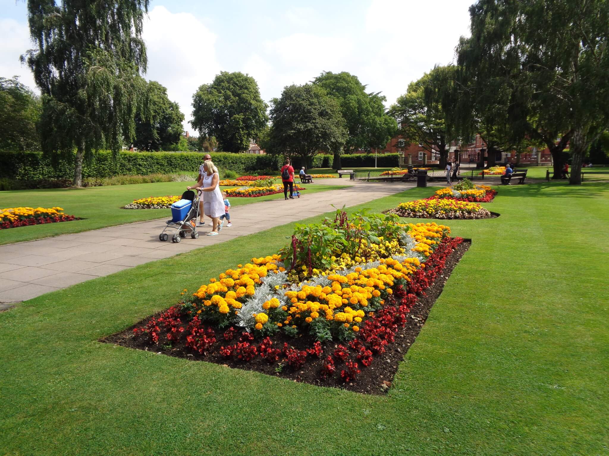A park with colorful flower beds, green lawns, and people walking, one pushing a stroller, on paved paths. - Home Instead