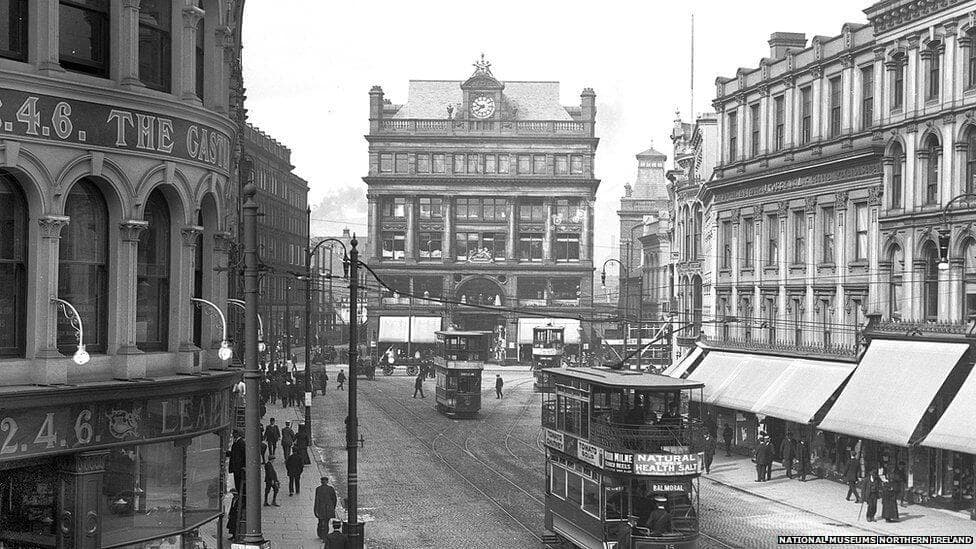 Black and white photo of a busy street with trams and pedestrians, featuring a large central building with a clock. - Home Instead