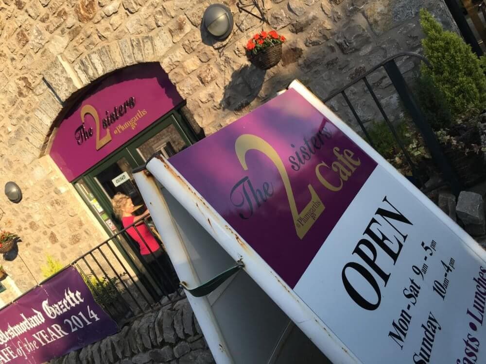 Sign for "The 2 Sisters Café" outside a stone building, with a smaller sign indicating it's open and operational hours. - Home Instead