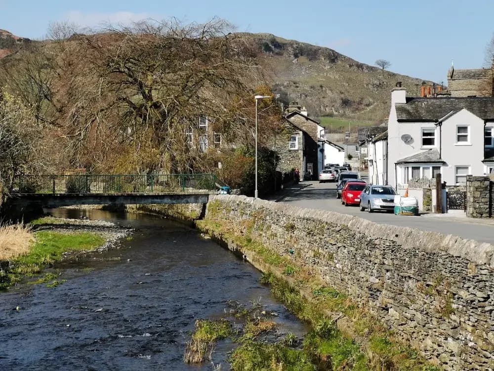 A small river runs through a quaint village with stone houses, a bridge, and cars parked along a narrow street. - Home Instead