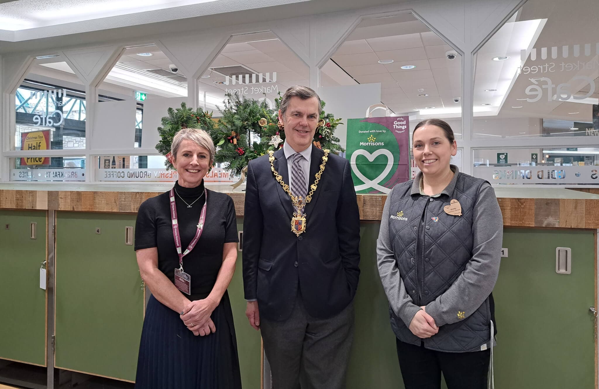 Three people standing indoors in front of a counter; one wearing a mayoral chain and the others in work uniforms. - Home Instead