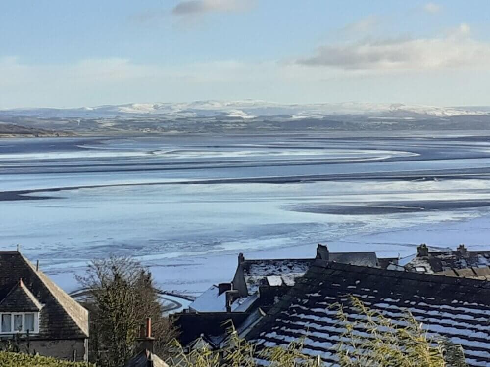 Snow-covered rooftops in the foreground, with a vast, icy river and distant, snow-capped mountains under a clear sky. - Home Instead