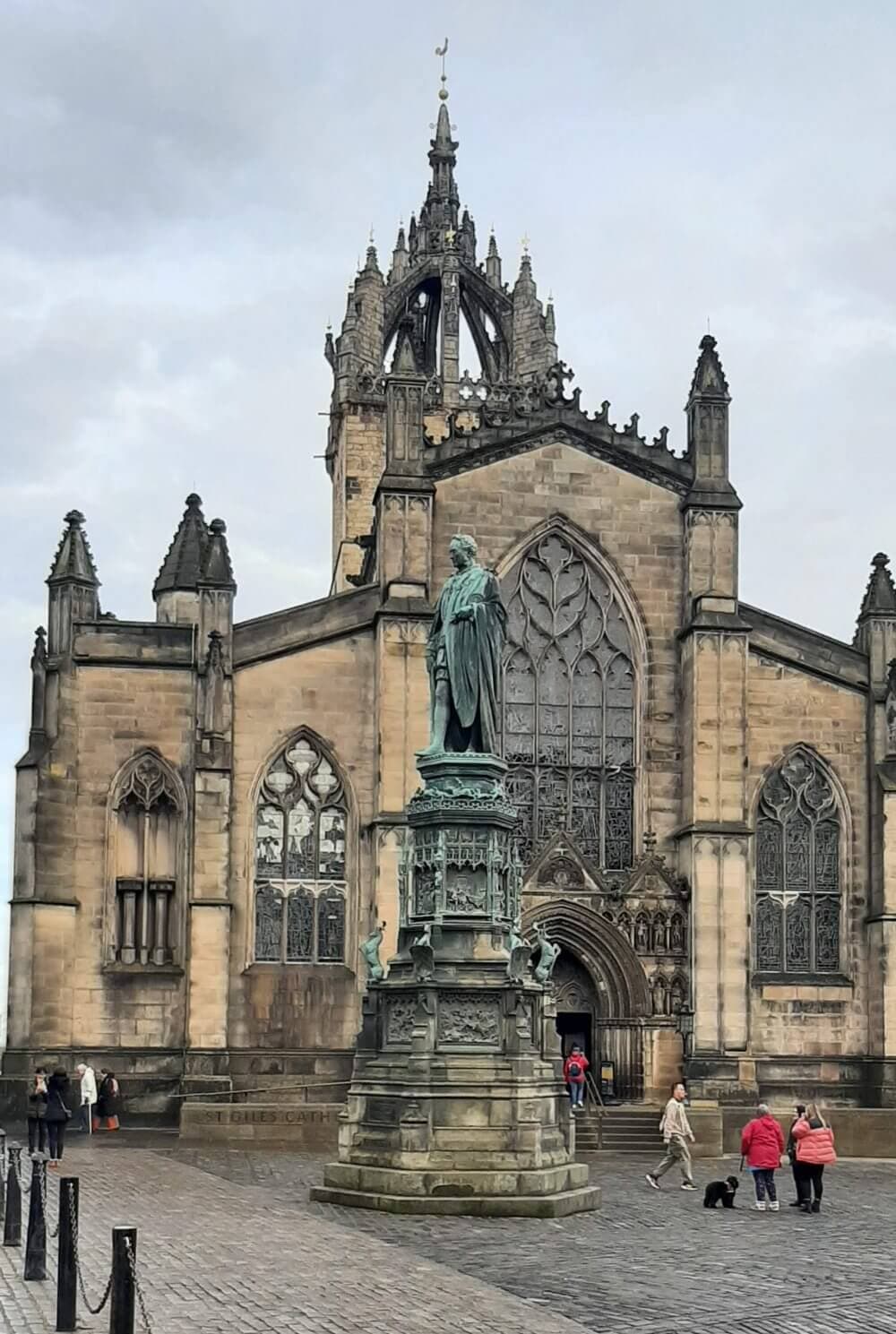 A historic cathedral with a statue in front, flanked by stone buildings under a cloudy sky, with people walking nearby. - Home Instead