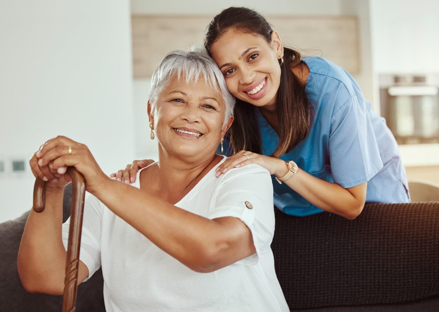 Elderly woman with a cane smiling with a younger woman in blue scrubs who has her hands on the older woman's shoulders. - Home Instead Bournemouth & Christchurch