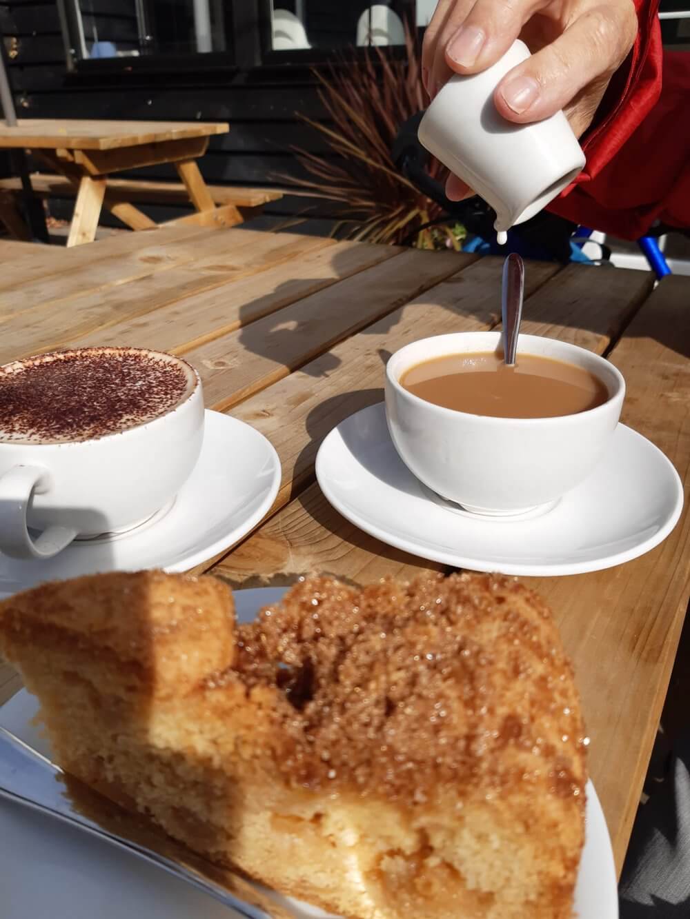 A hand pours milk into a cup of tea on a wooden table. Nearby are a cappuccino and a slice of cake. - Home Instead