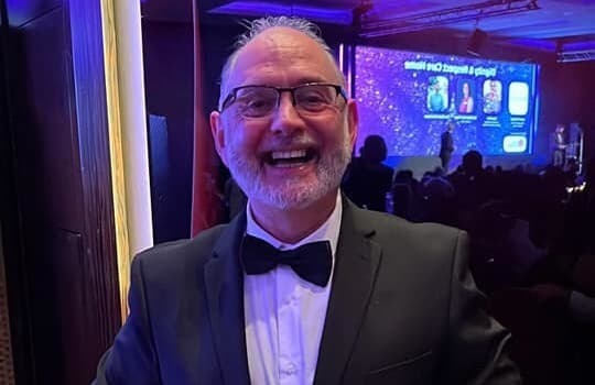 A man wearing glasses and a tuxedo smiles at an indoor event with blurred screen and audience in the background. - Home Instead