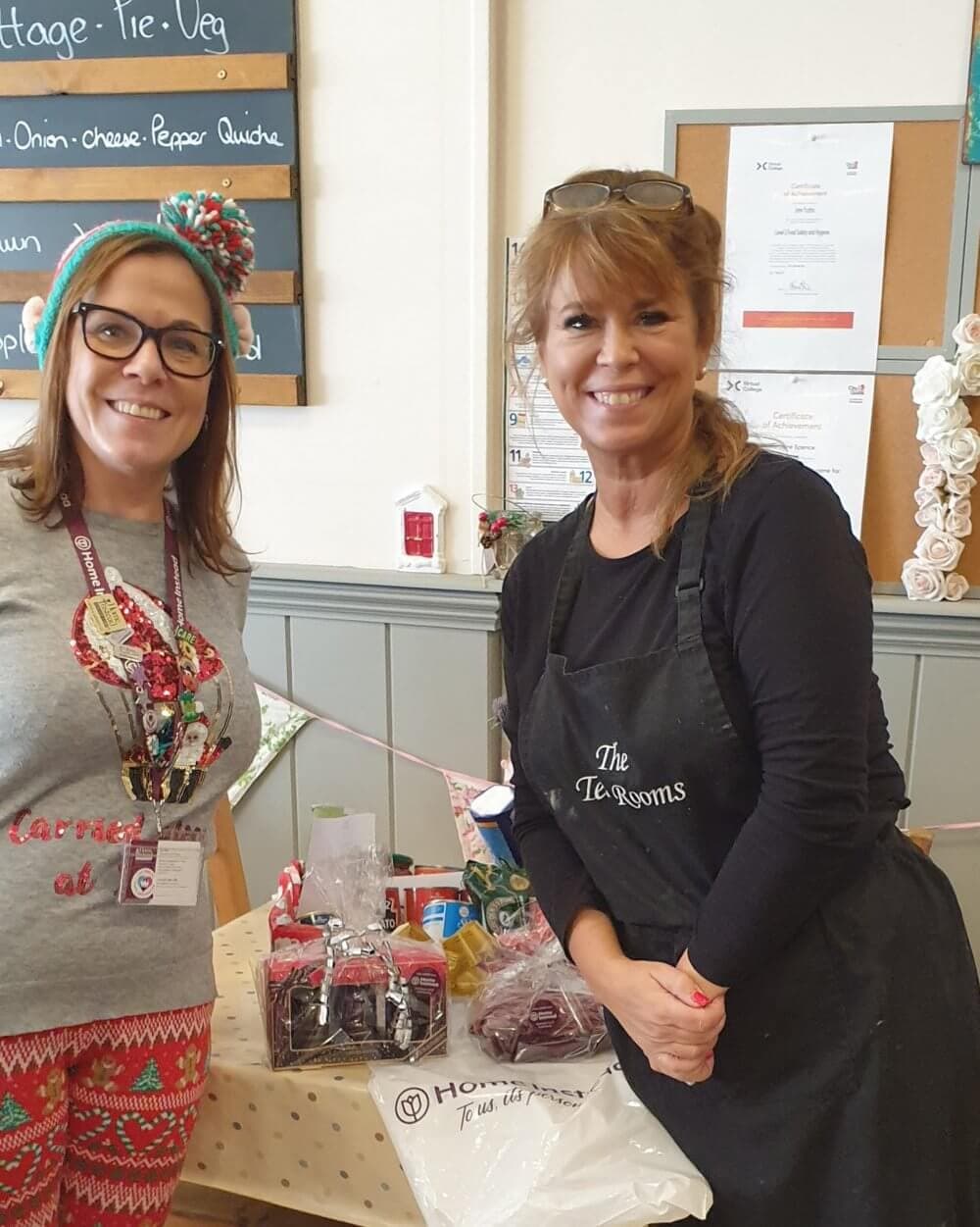 Two women smiling and standing by a table with gift items in a tea room. A menu board is visible in the background. - Home Instead