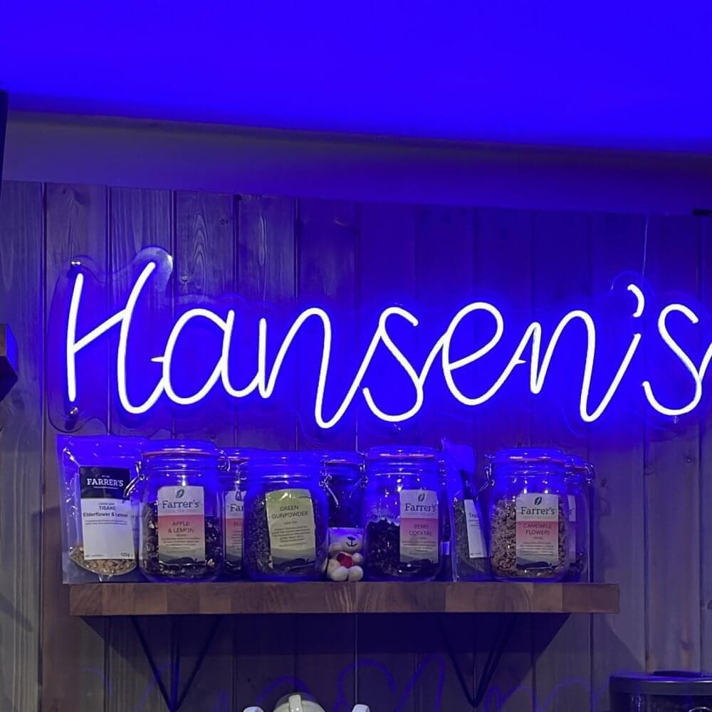 A neon sign reading "Hansen's" is displayed above a wooden shelf holding jars and bags of assorted goods. - Home Instead