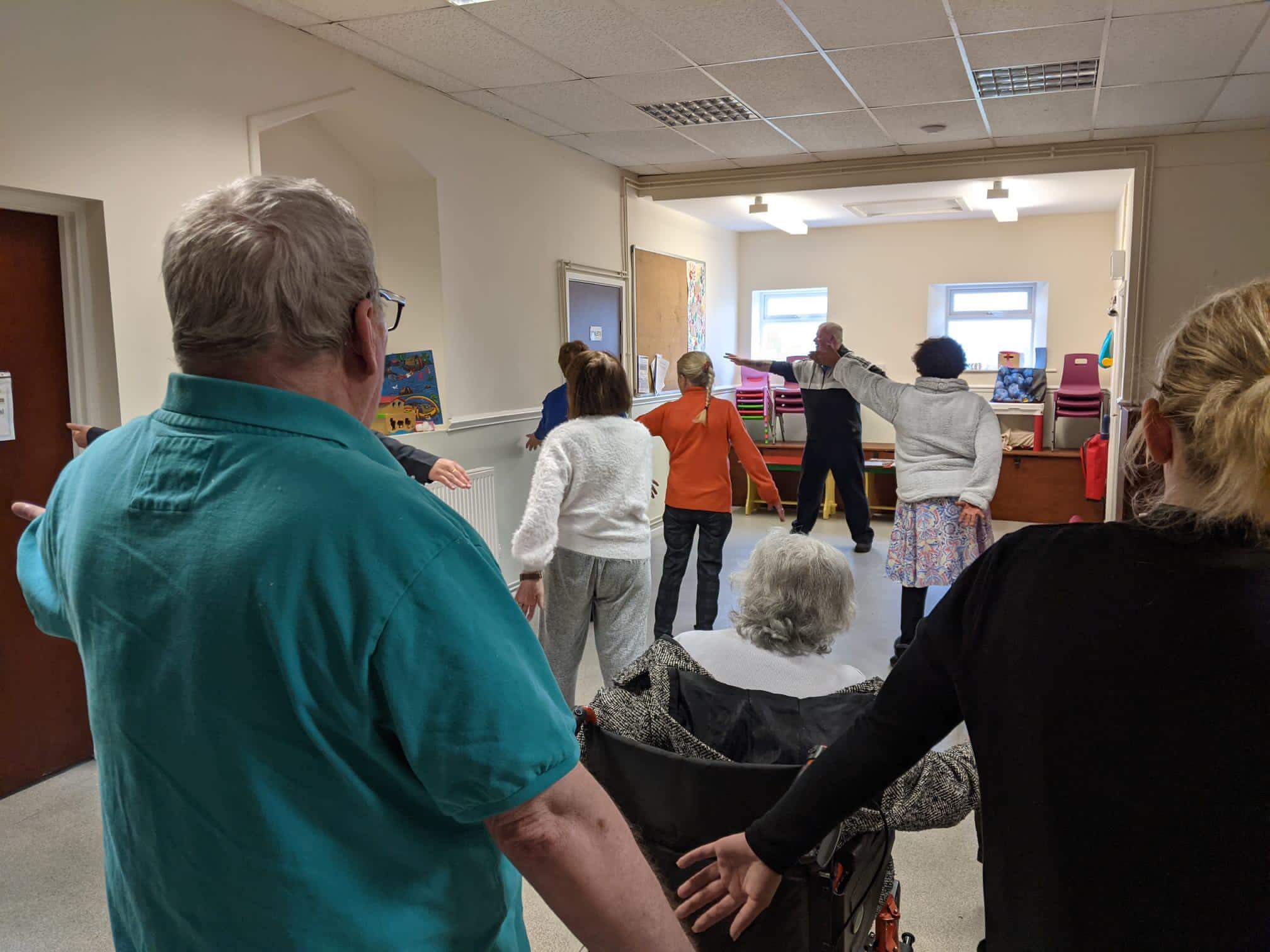 A group of adults and a person in a wheelchair participate in an exercise class, performing stretches with arms extended. - Home Instead
