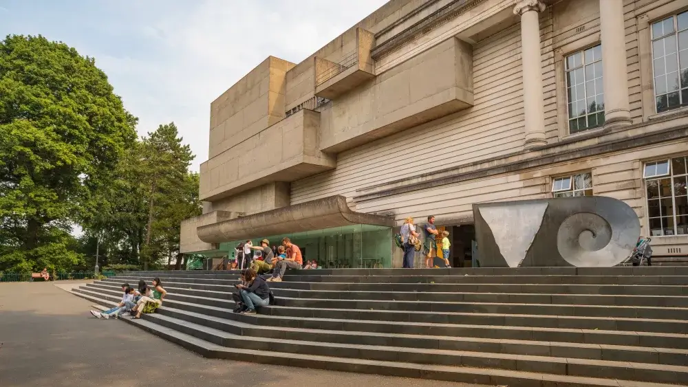 People sitting on stairs outside a modern building with a large sculpture and trees in the background. - Home Instead