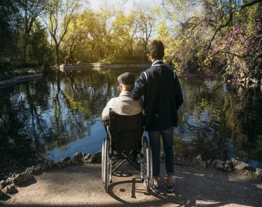 A person stands beside a wheelchair user, overlooking a peaceful pond in a tree-filled park backlit by the sun. - Home Instead