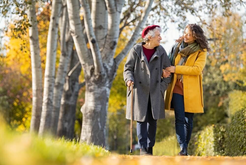 An elderly woman with a cane and a younger woman walk arm in arm on a tree-lined path during autumn. - Home Instead
