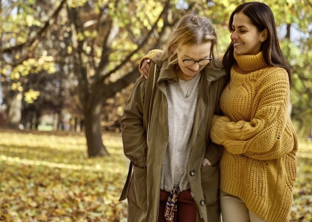 Two women walking closely in a park with autumn leaves, one wearing a green coat and the other in a yellow sweater. - Home Instead