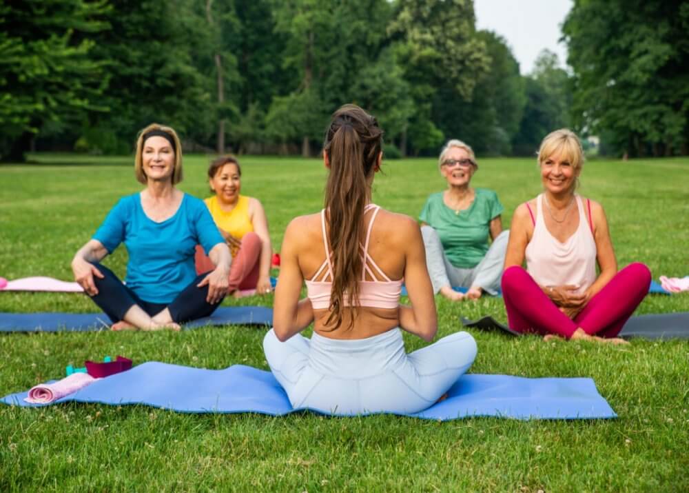 A group of women practicing yoga outdoors on a grassy field, led by a woman facing them with her back to the camera. - Home Instead