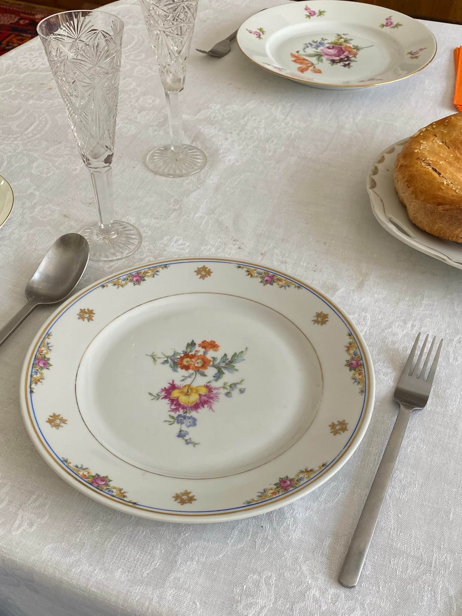 Elegant table setting with floral porcelain plates, crystal glasses, a spoon and fork, and a pastry on a white tablecloth. - Home Instead