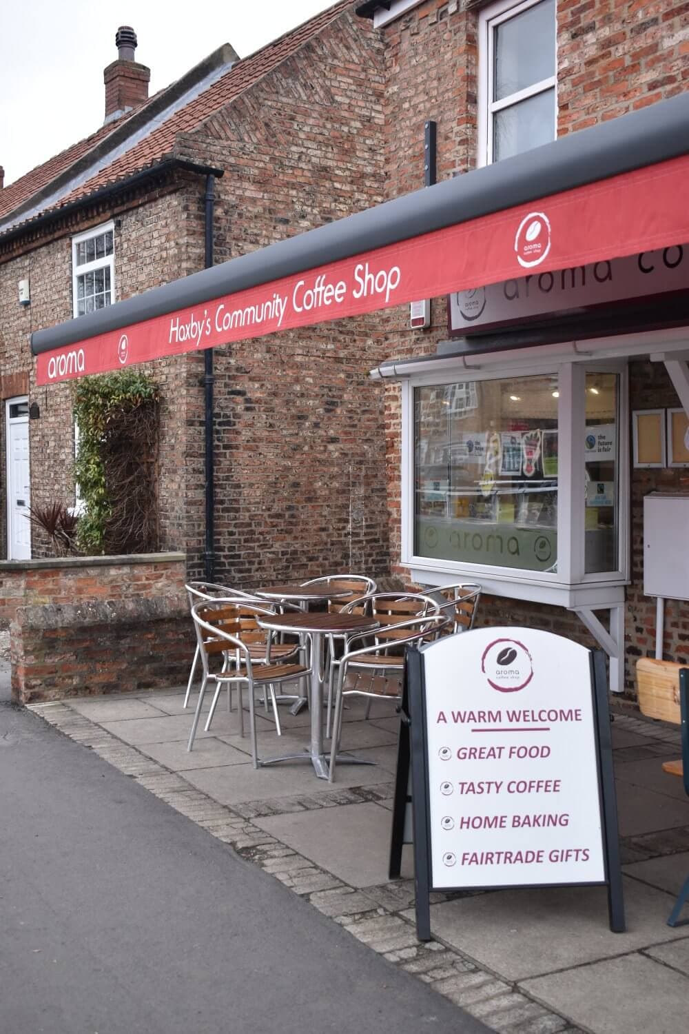 Exterior of a brick coffee shop with outdoor seating and a signboard, under an awning reading "Haxby's Community Coffee Shop. - Home Instead