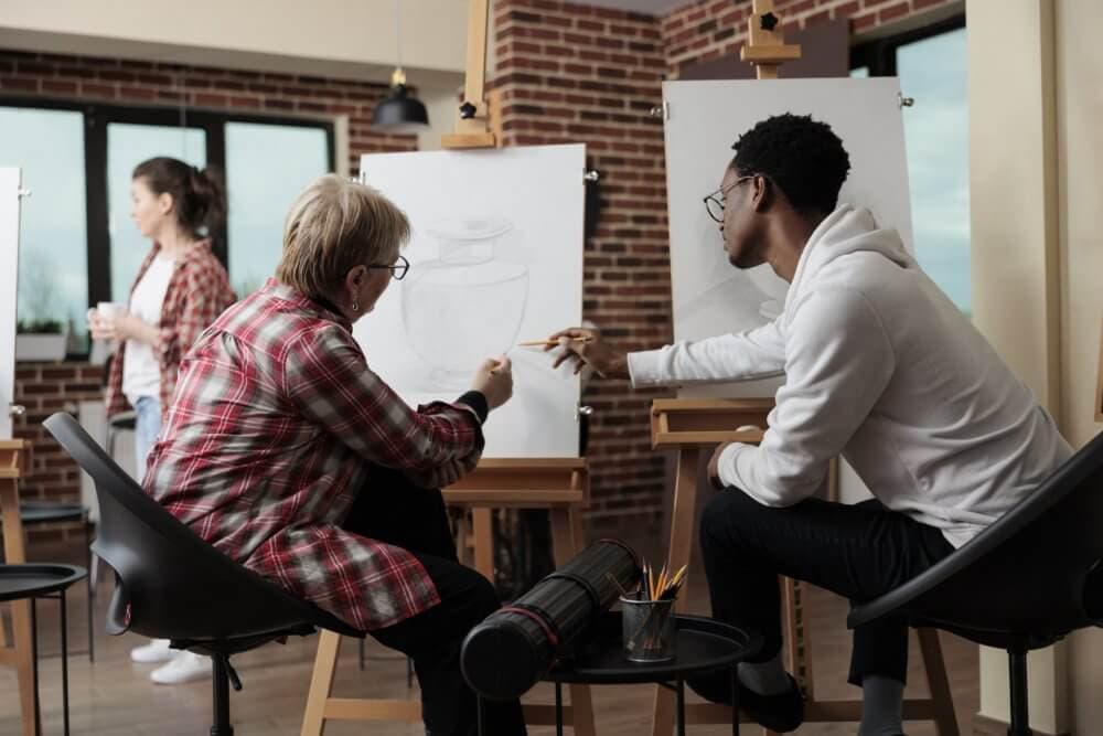 Two people in an art studio, one in a plaid shirt, another in a white hoodie, discussing sketches on easels. - Home Instead