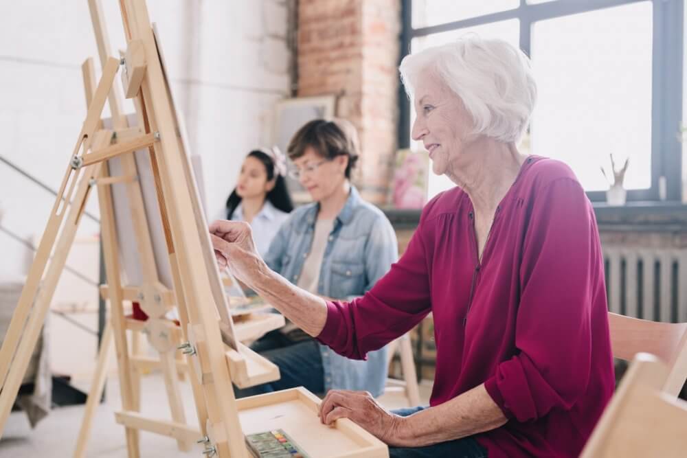 Three women of different ages painting on canvases in a bright art studio with wooden easels and large windows. - Home Instead