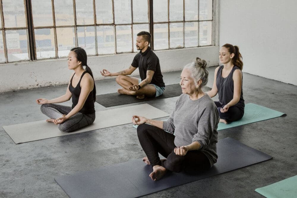 A diverse group of people sits in lotus position on yoga mats in a bright, industrial space with large windows. - Home Instead