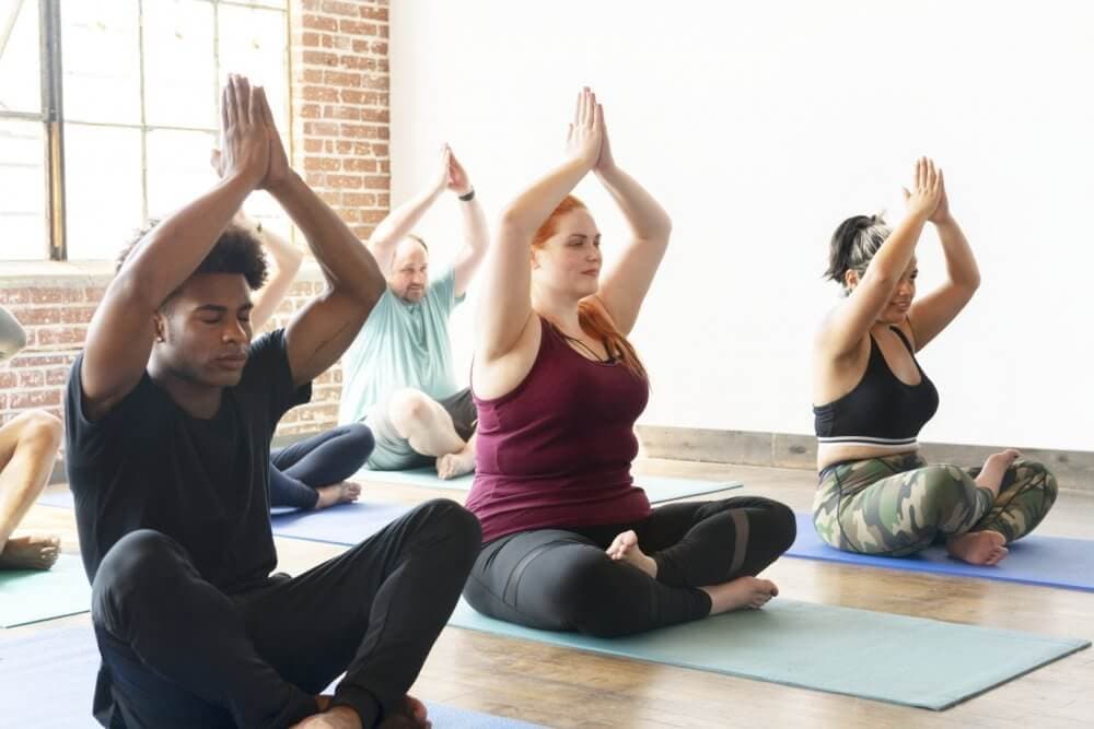 A group of people sits cross-legged on yoga mats in a studio, practicing a pose with hands held together above their heads. - Home Instead Bournemouth & Christchurch