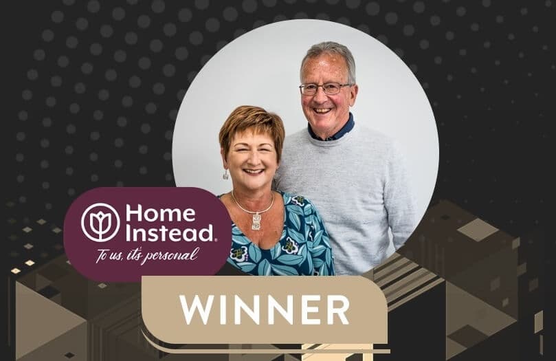 A smiling couple stands together in front of a "Home Instead" logo with the word "WINNER" beneath them. - Home Instead