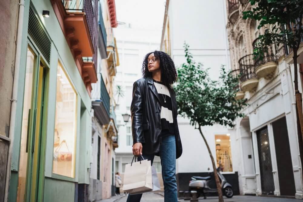 Person with curly hair wearing a leather jacket, holding a shopping bag, walks down a narrow street lined with buildings. - Home Instead
