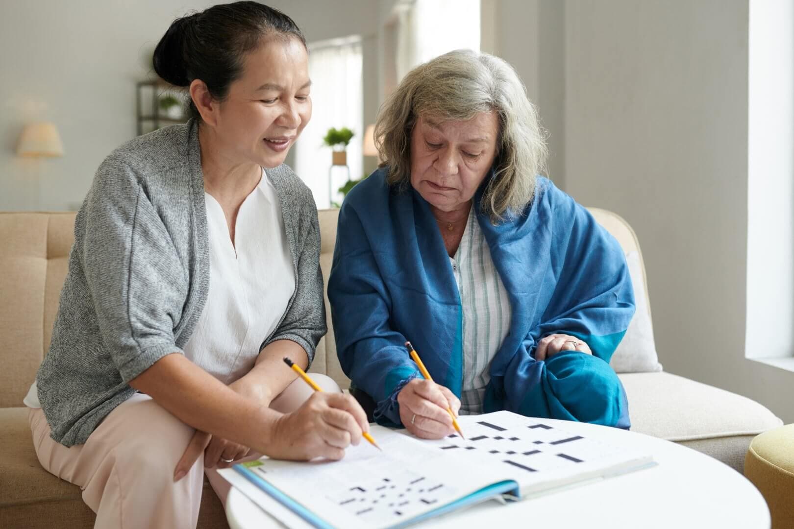 Two women sitting on a couch, working on a crossword puzzle together, both holding pencils. - Home Instead