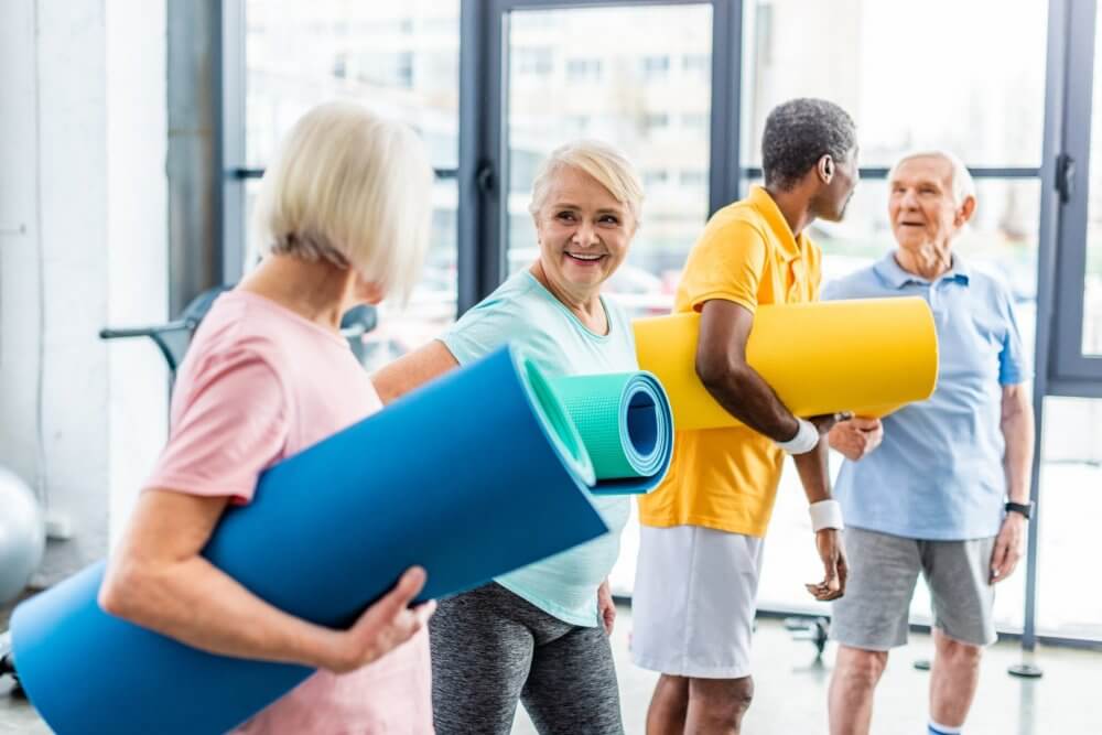 Group of seniors at a fitness class smiling and holding yoga mats, ready for their workout session. - Home Instead