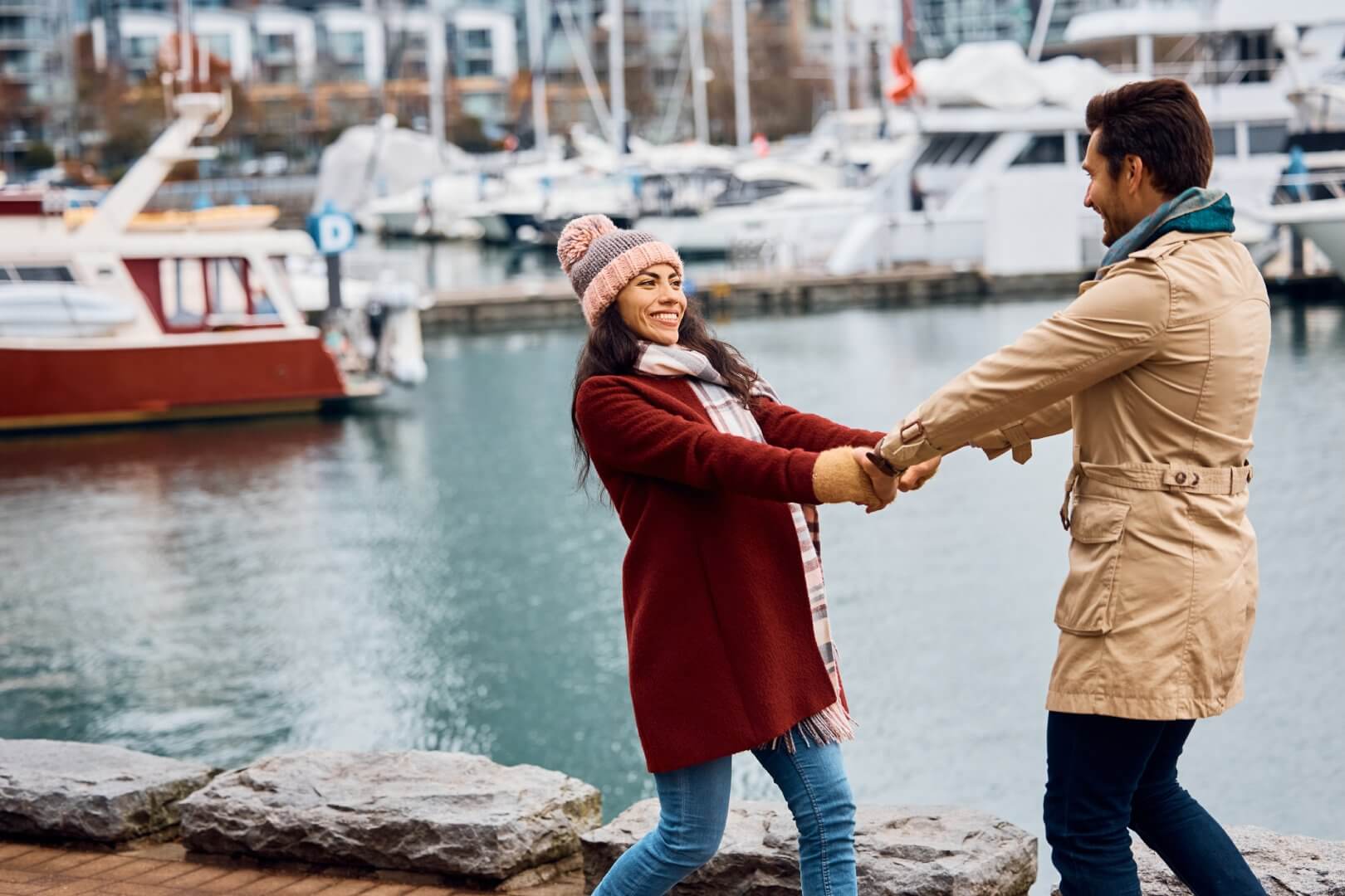 Two people joyfully holding hands near a marina, clad in winter clothing with boats and water in the background. - Home Instead Bournemouth & Christchurch