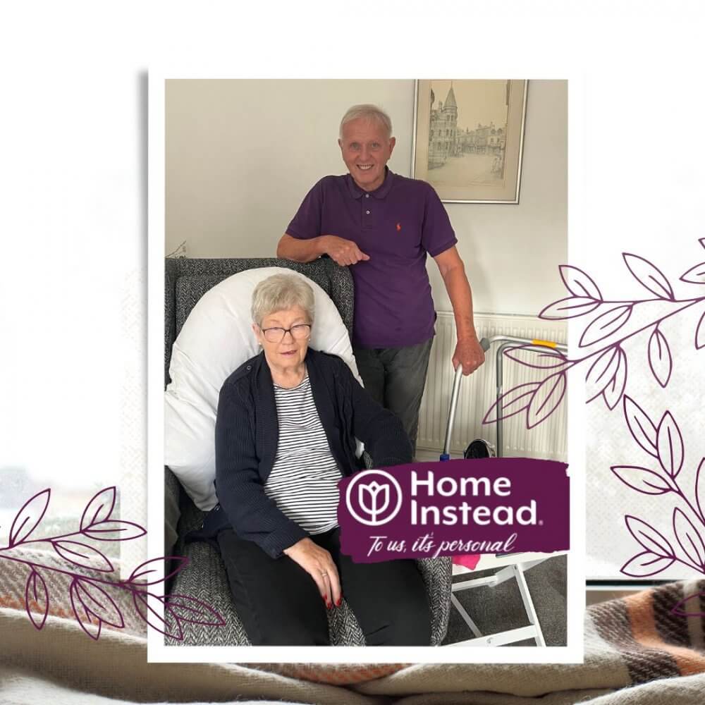 A senior woman sits in a chair with a man standing beside her, both smiling with a "Home Instead" logo in the foreground. - Home Instead