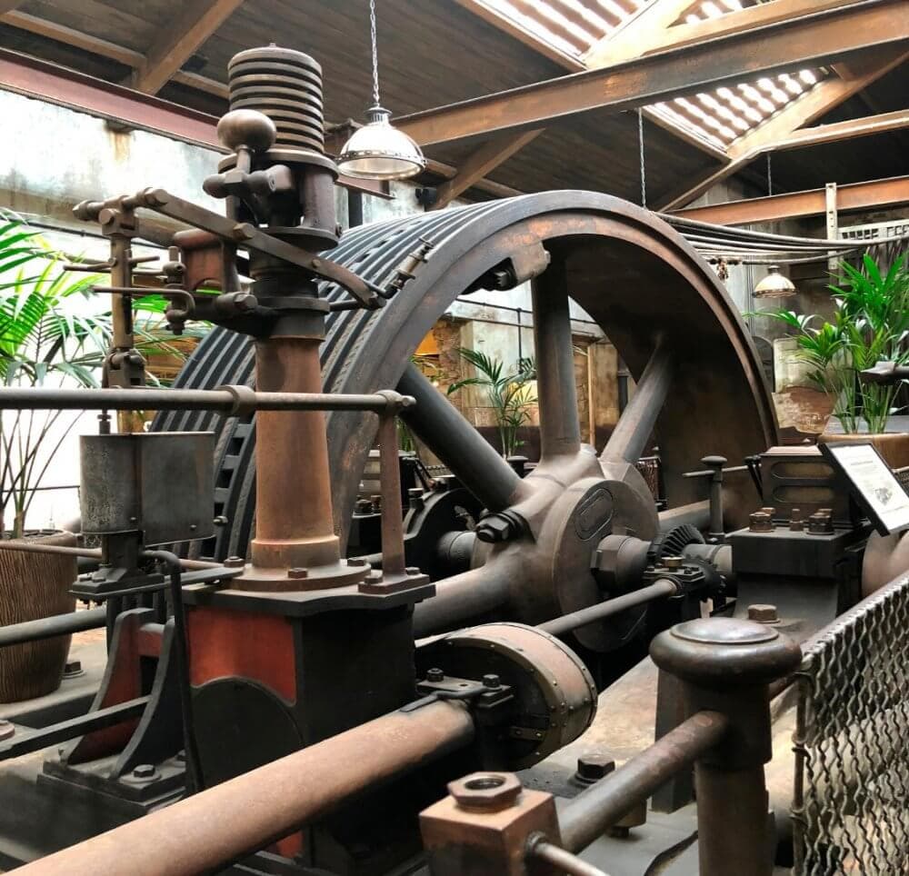 A large, historic industrial machine with gears and levers, displayed in a well-lit, indoor setting with plants nearby. - Home Instead