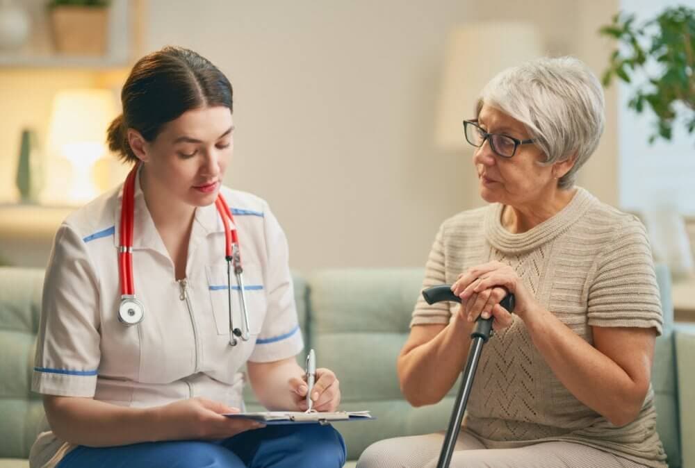 A nurse sits and writes on a clipboard while talking to an elderly woman with glasses and a walking cane. - Home Instead Bournemouth & Christchurch