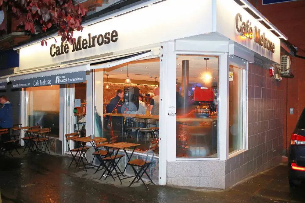 Exterior of Café Melrose on a rainy evening, with people inside and empty tables and chairs outside. - Home Instead